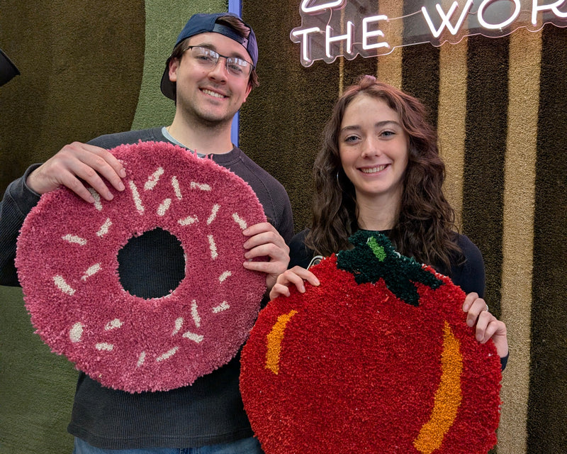 Two women standing in front of a 'TUF THE WORLD' sign, holding up hand-pile-rupee character rugs. The woman on the left holds a Toad-themed rug, and the woman on the right holds a yellow smiley face rug.