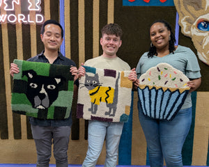 Three people standing indoors holding handmade tufted rugs with an animal and cupcake-shaped rugs. The rug on the left is green and black with a cat face, the center rug is cream and yellow with a cat, and the right rug is blue and white with a cupcake.